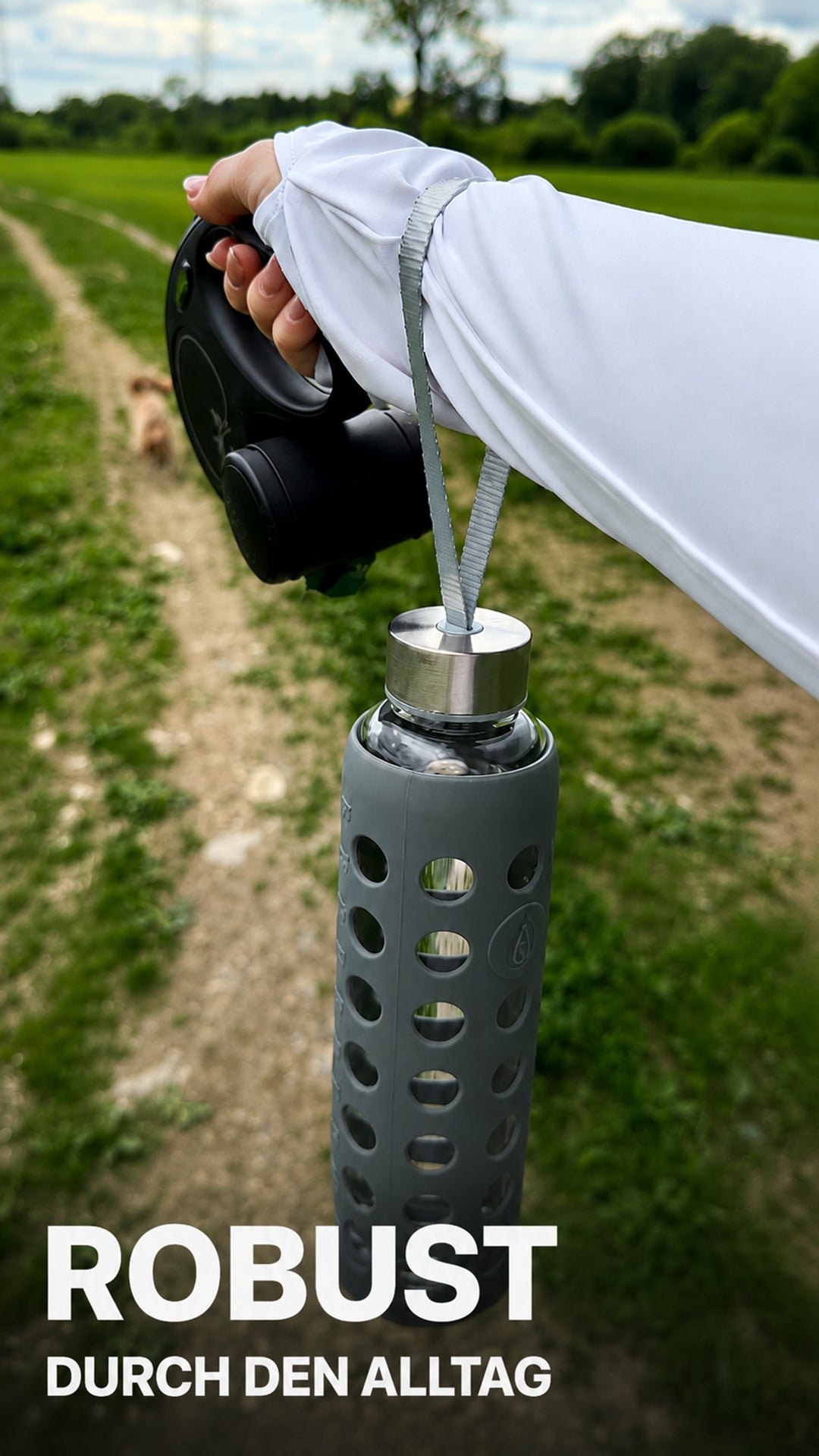 Load video: Eine junge Frau zeigt auf einem Feldweg eine spottle Glasflasche mit grauer Silikonhülle und Trageschlaufe und berichtet, warum sie die Flasche als robust und sicher im Alltag empfindet. 