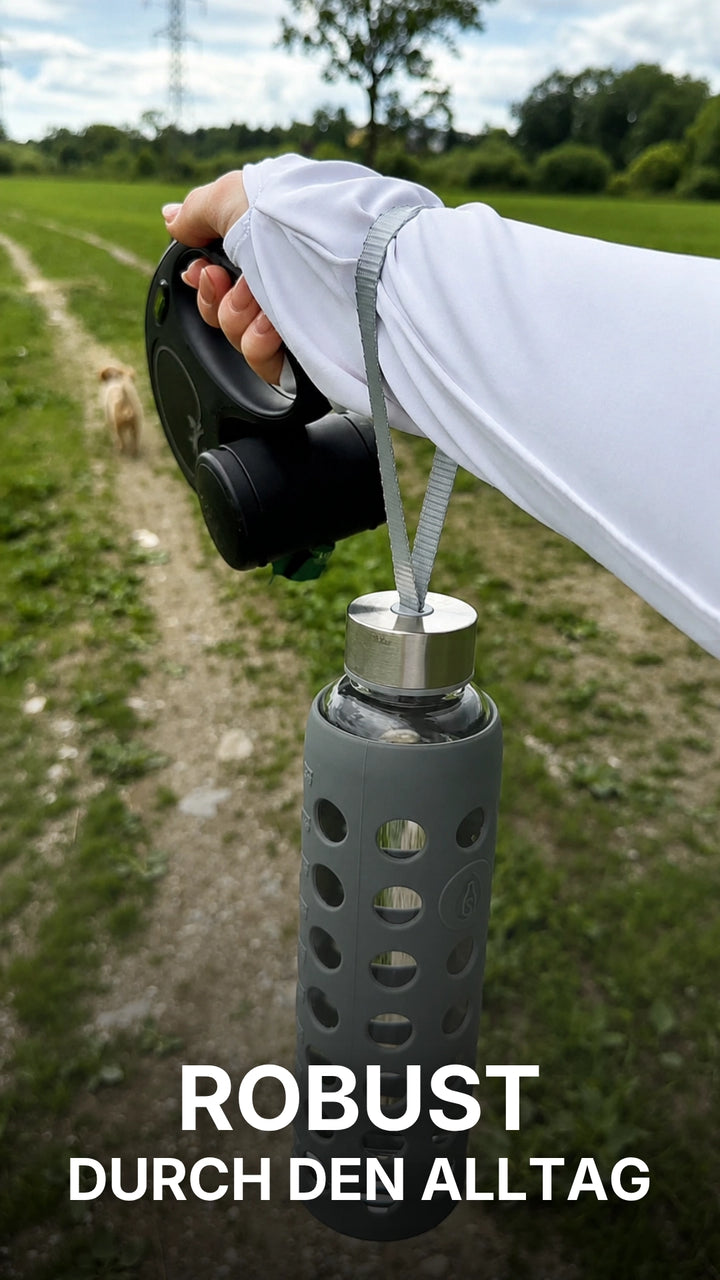 Eine junge Frau zeigt auf einem Feldweg eine spottle Glasflasche mit grauer Silikonhülle und Trageschlaufe und berichtet, warum sie die Flasche als robust und sicher im Alltag empfindet. 