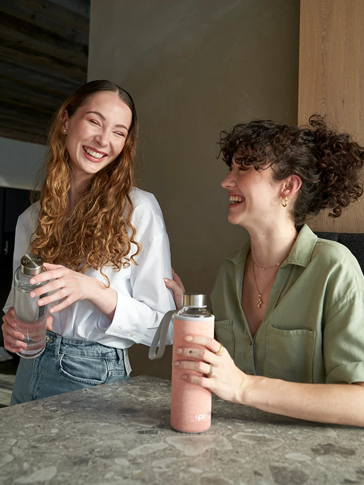 2 Mädchen, mit Trinkflaschen aus Glas, sitzen lächelt am Küchentisch.