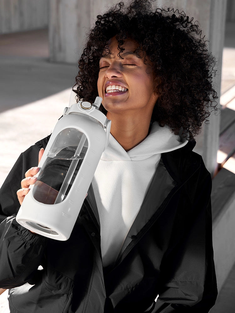 Ein Mädchen sitzt auf einer Bank und genießt einen Schluck aus einer großen Trinkflasche mit 2 Litern Fassungsvermögen. Dabei lächelt sie über das ganze Gesicht.